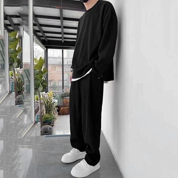 A person wearing a loose black long-sleeve top and black pants paired with white sneakers, standing indoors next to a white wall.