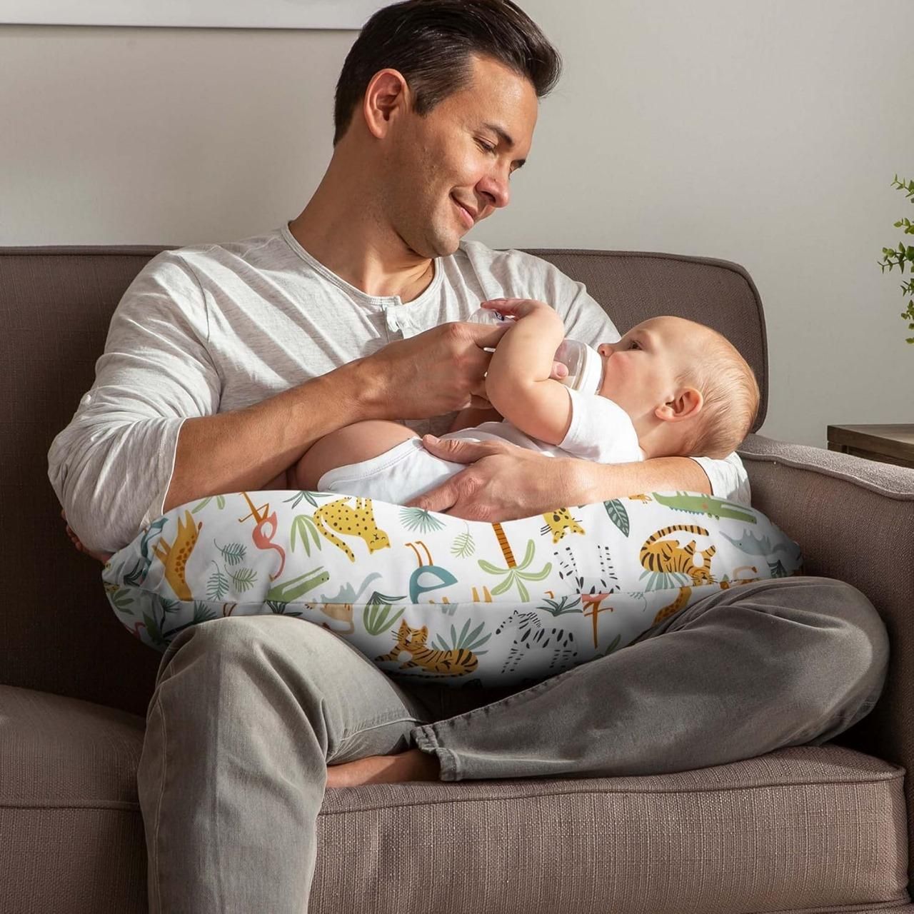 A man sitting on a couch feeding a baby with a bottle, using a colorful pillow with animal prints for support.