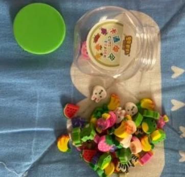 An open plastic container with a green lid, spilling out colorful erasers shaped like fruit and animals onto a blue surface.