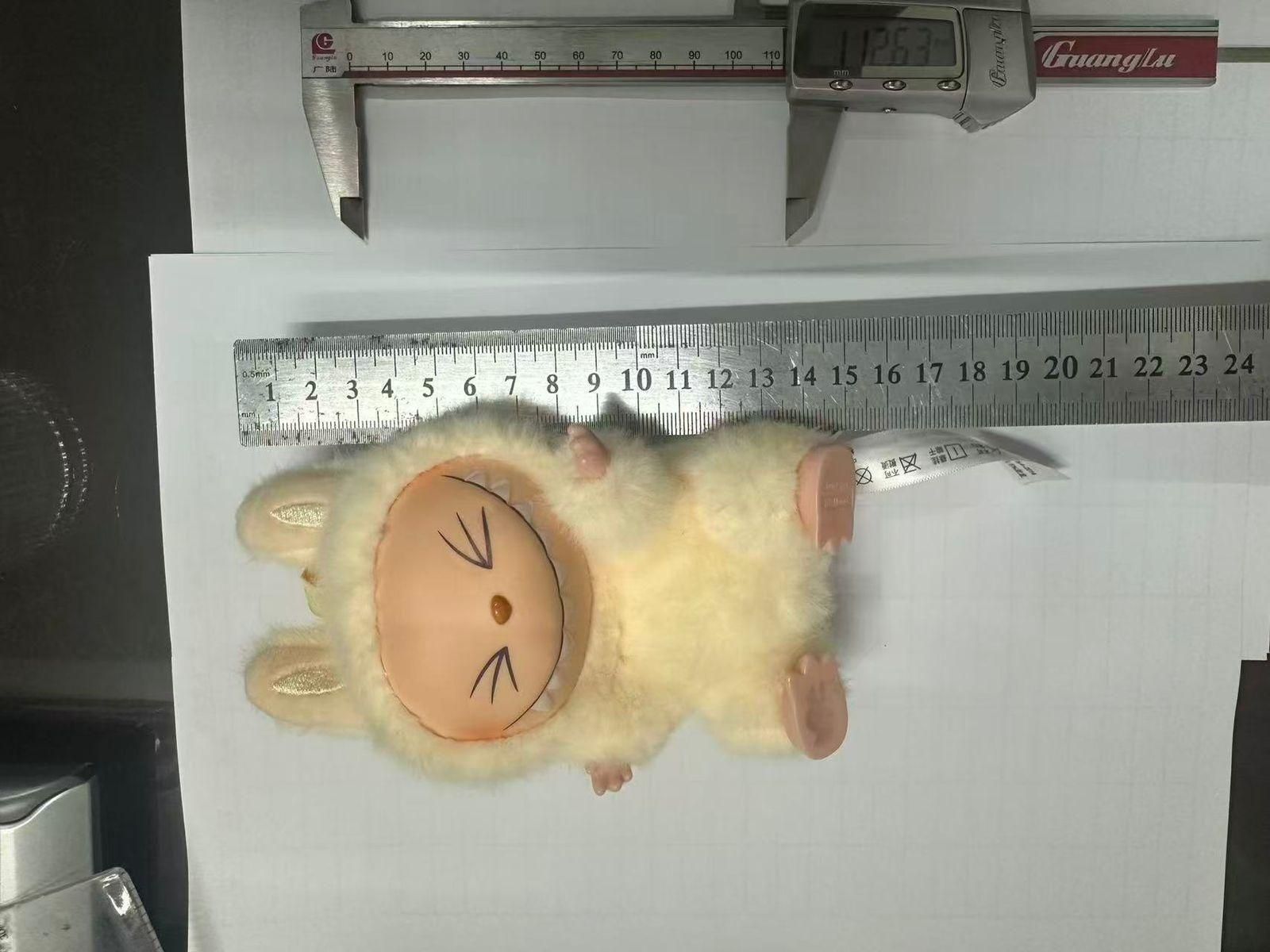 A plush toy with a round face and closed eyes, dressed in a fuzzy cream costume with bunny ears, lying next to a metal ruler and digital caliper for measurement.