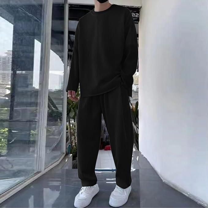 A person wearing a loose-fitting black long-sleeve shirt and black pants with white sneakers, standing indoors near large windows.