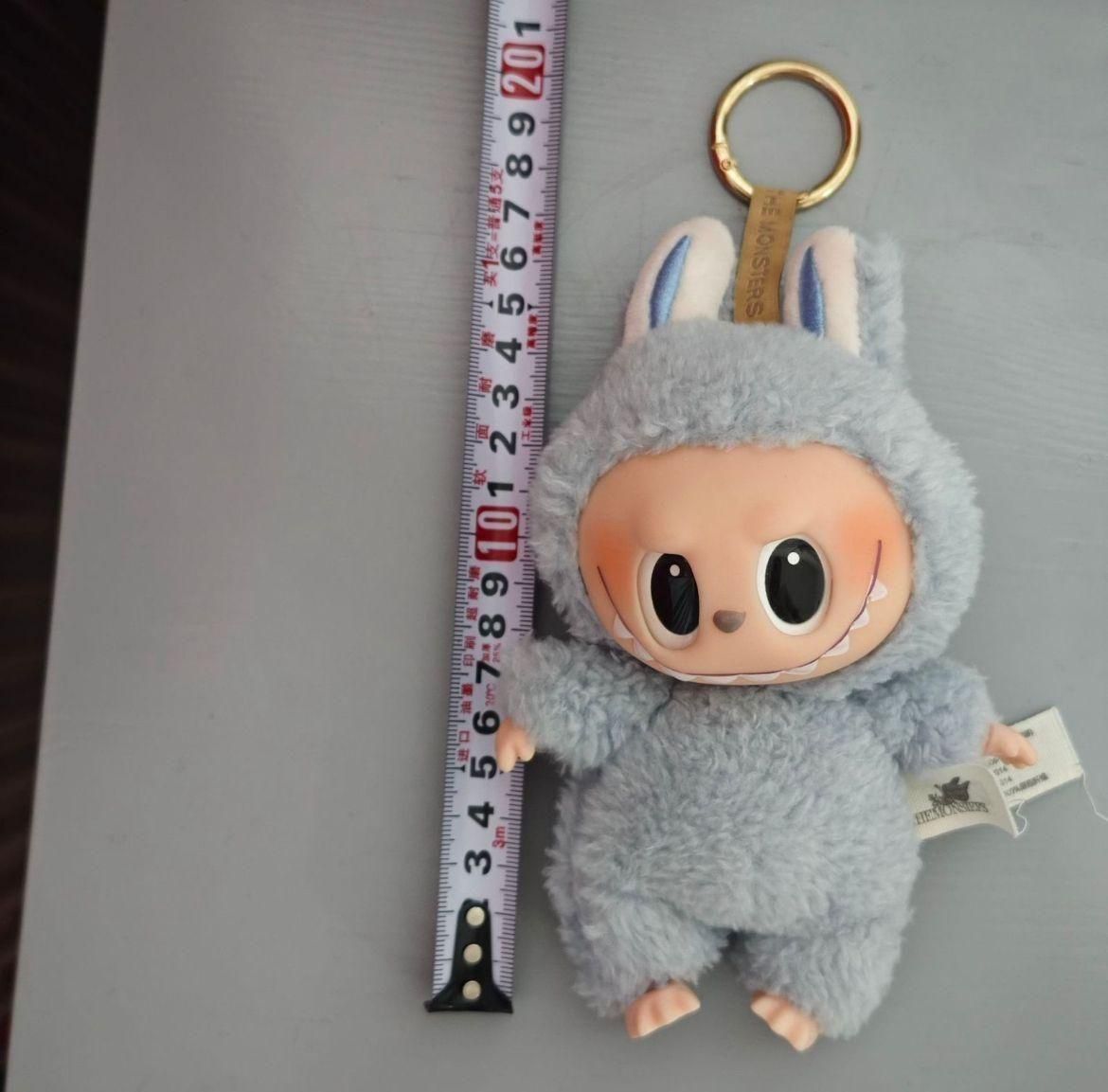 A plush keychain doll with big eyes and a gray furry bunny suit lying next to a measuring tape.