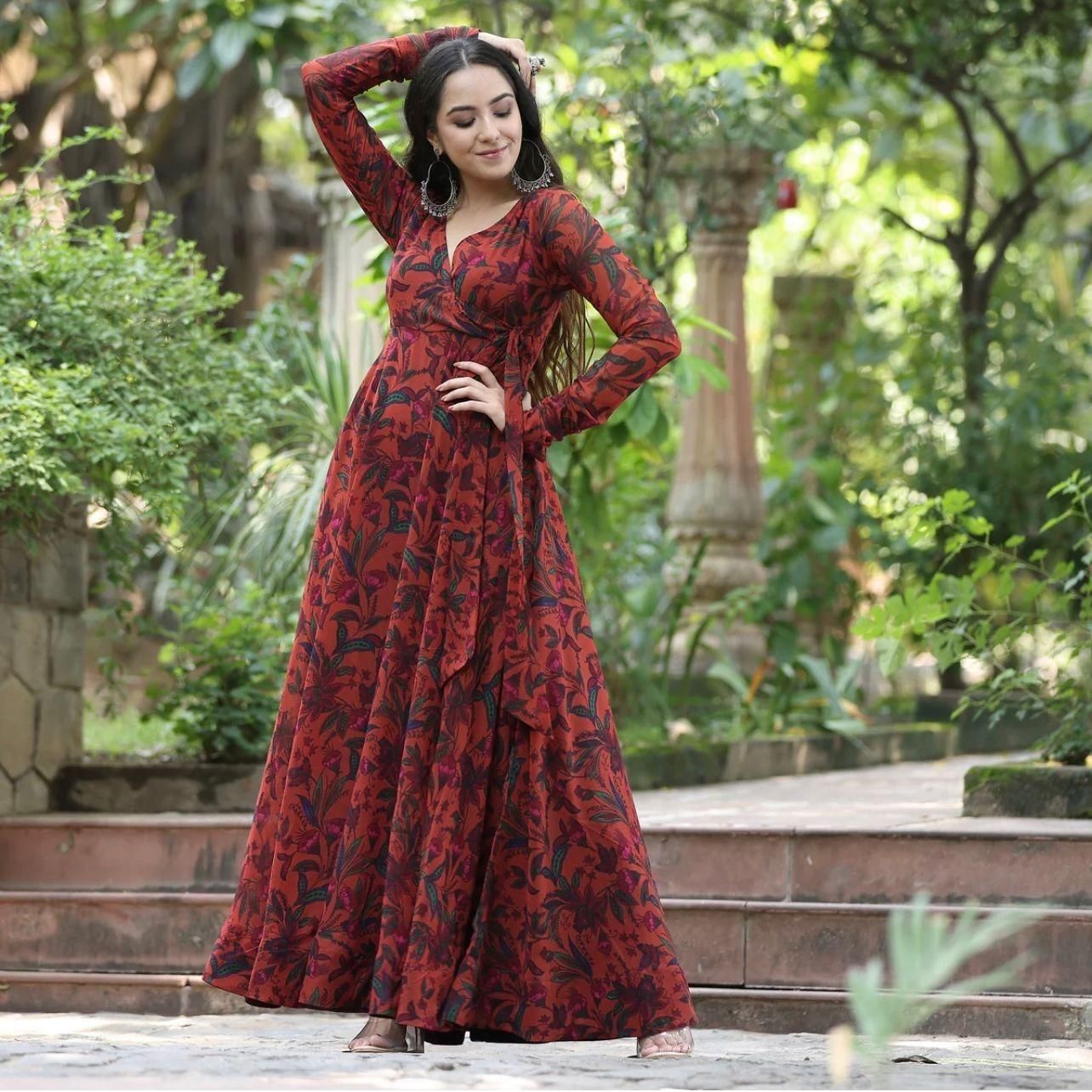 A woman wearing a long, flowing red and black floral dress posing outdoors on stone steps surrounded by greenery.