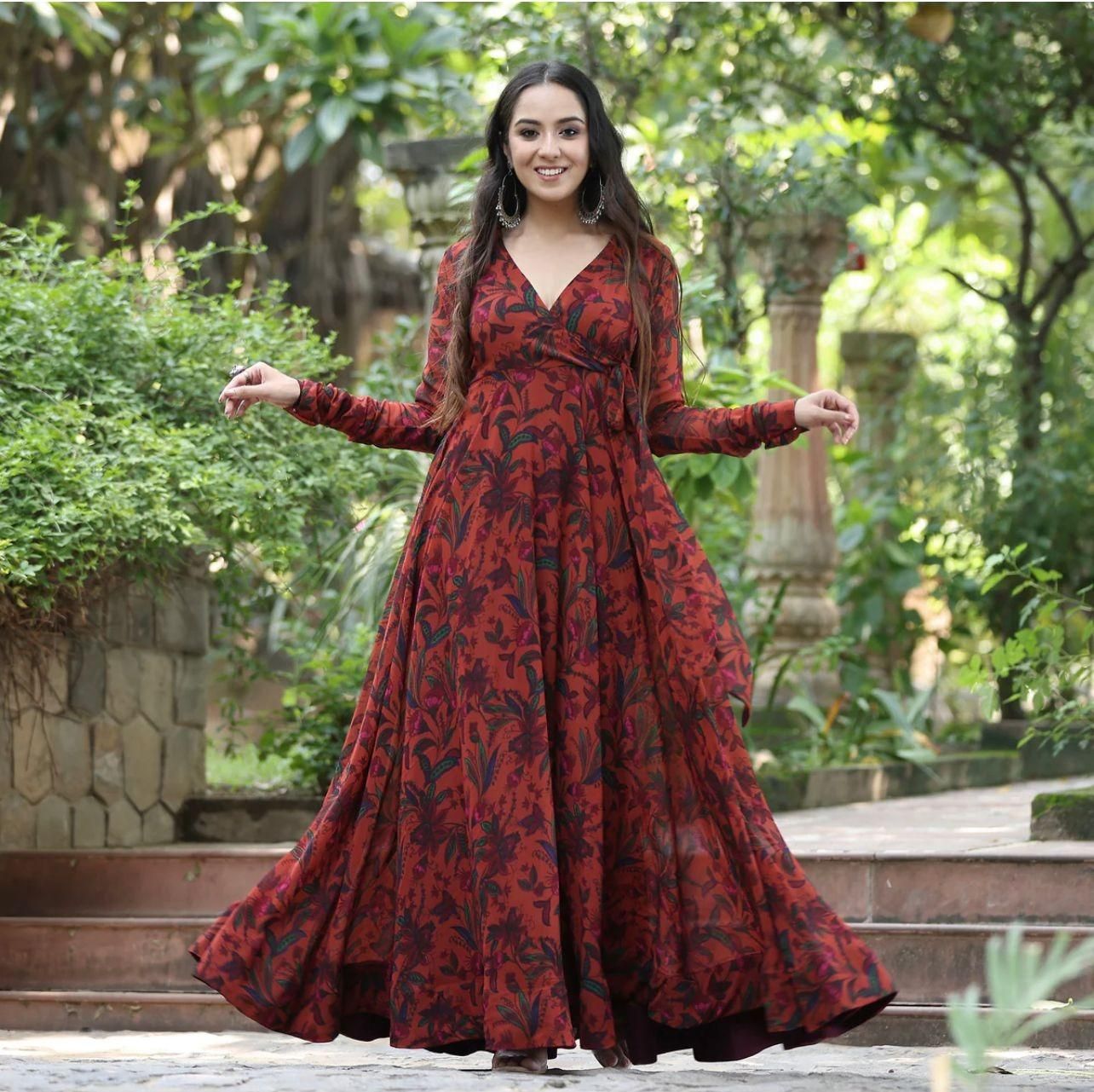 A woman wearing a long, flowing red and black floral dress standing outdoors surrounded by greenery.