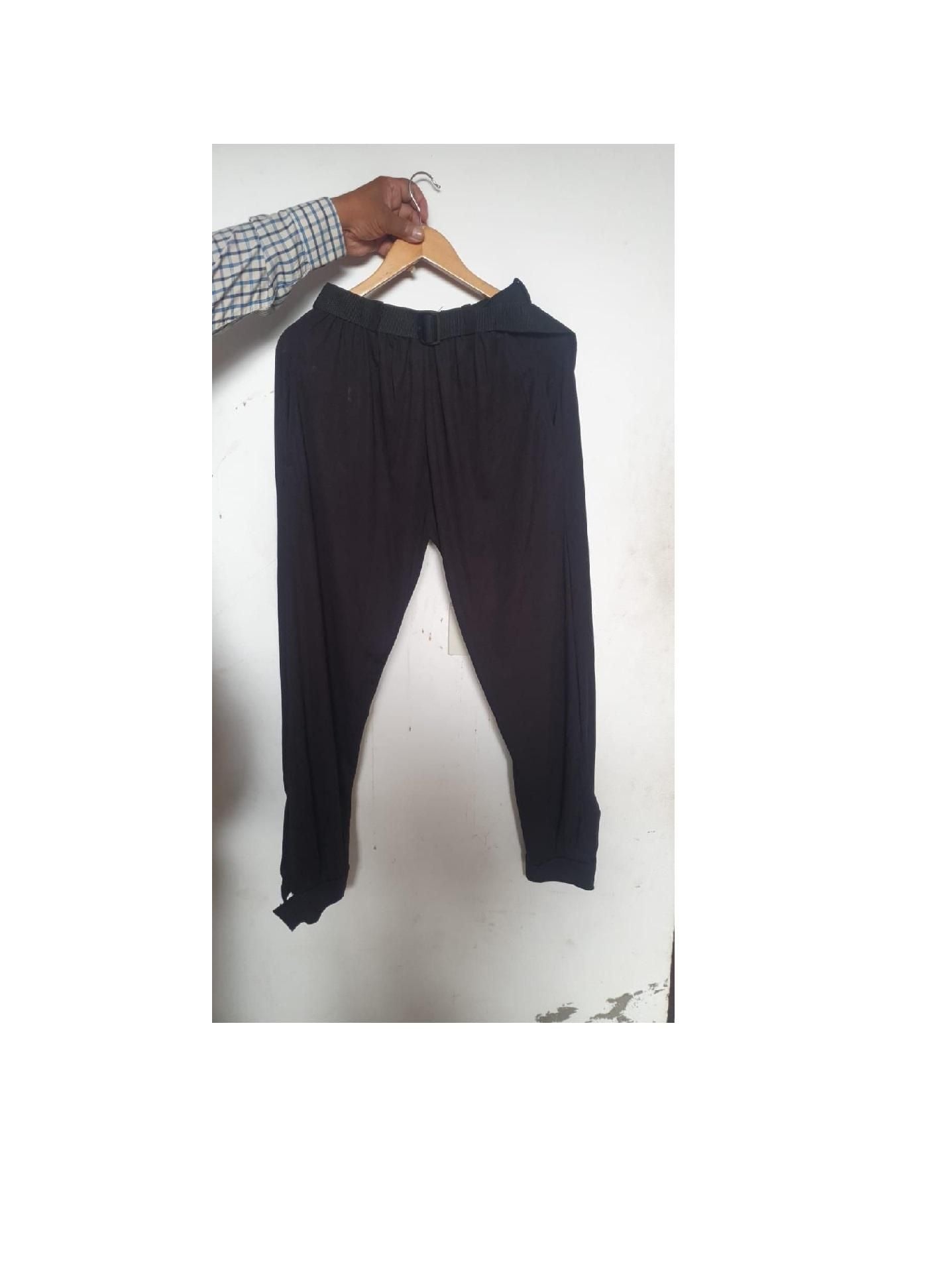 A pair of black pants hanging on a wooden hanger, held by a person wearing a checkered shirt.