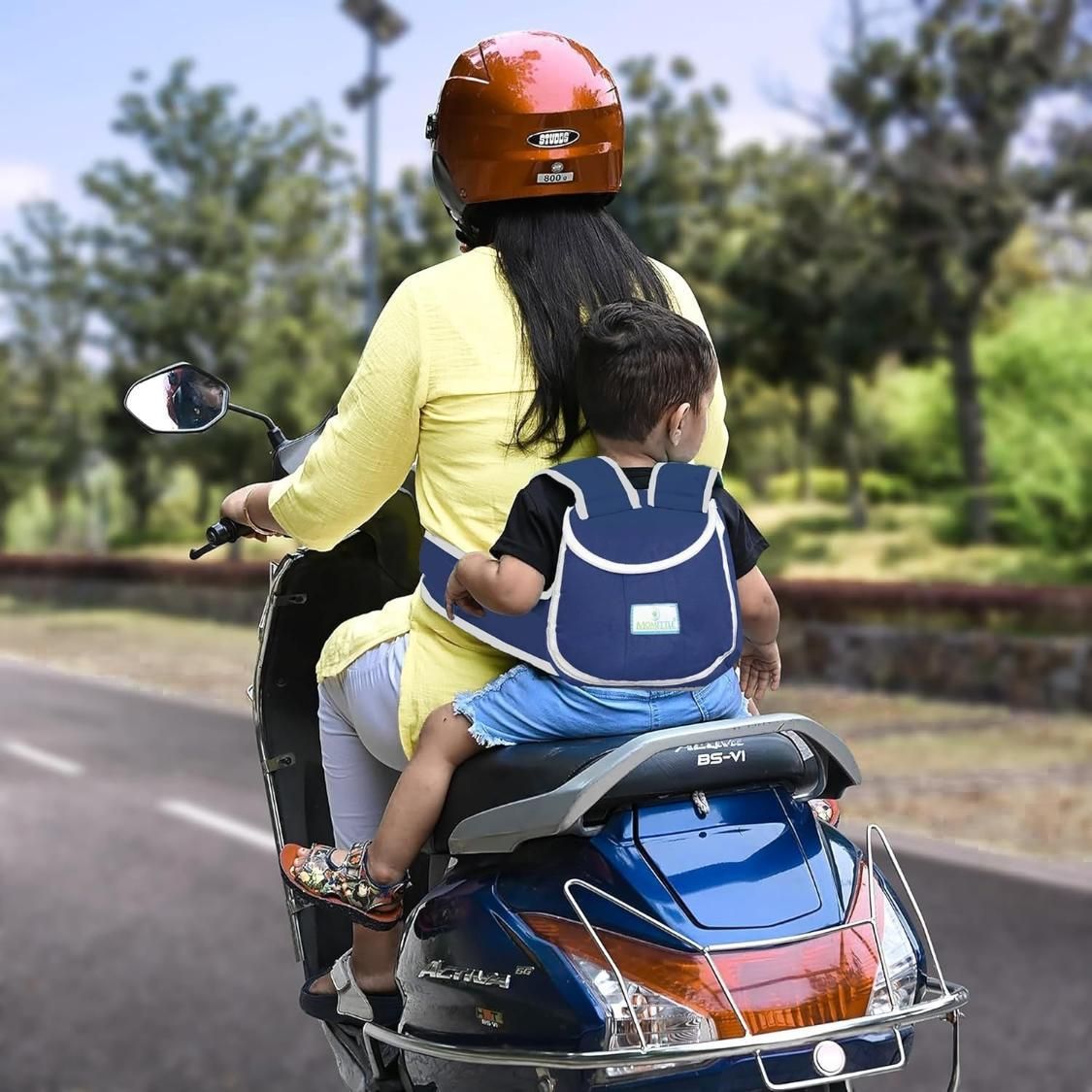 An adult wearing a helmet rides a blue scooter on a road with a young child secured in a safety harness on the back seat.