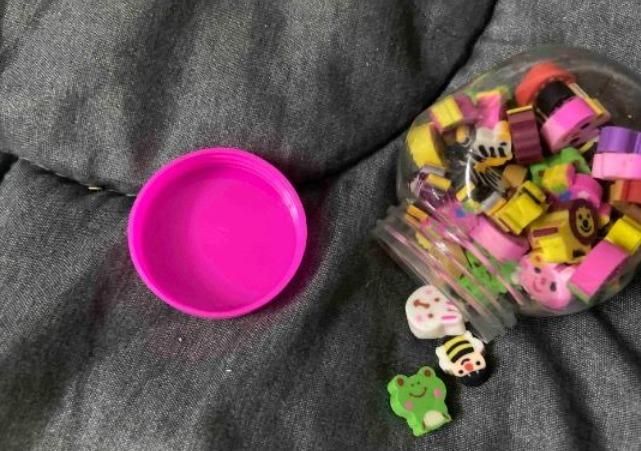 A plastic container with colorful erasers shaped like animals and objects, some spilled out on a gray fabric surface.