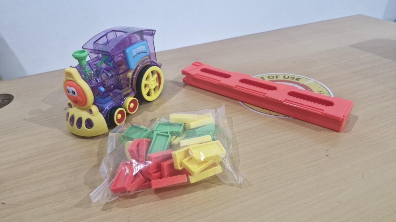 A colorful toy train with a transparent purple body, yellow wheels, a red toy track piece, and a plastic bag containing red, yellow, and green plastic blocks on a wooden surface.