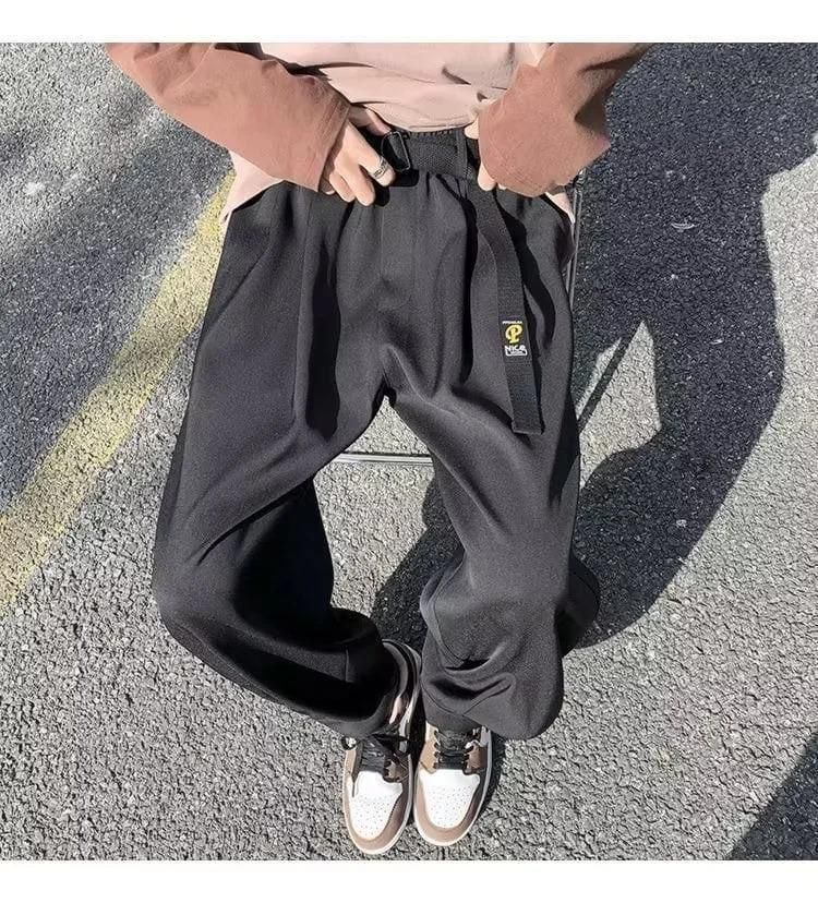 A person wearing loose black pants, a brown long-sleeve shirt, and two-tone sneakers sitting on a chair outdoors.