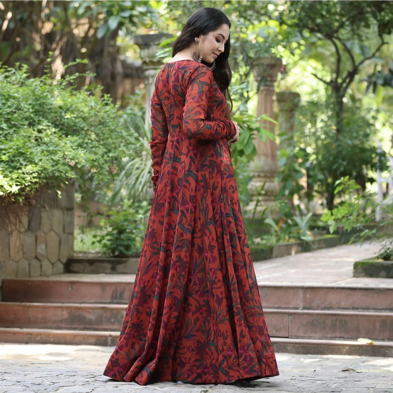 A woman in a long, red floral dress standing outdoors in a lush garden.