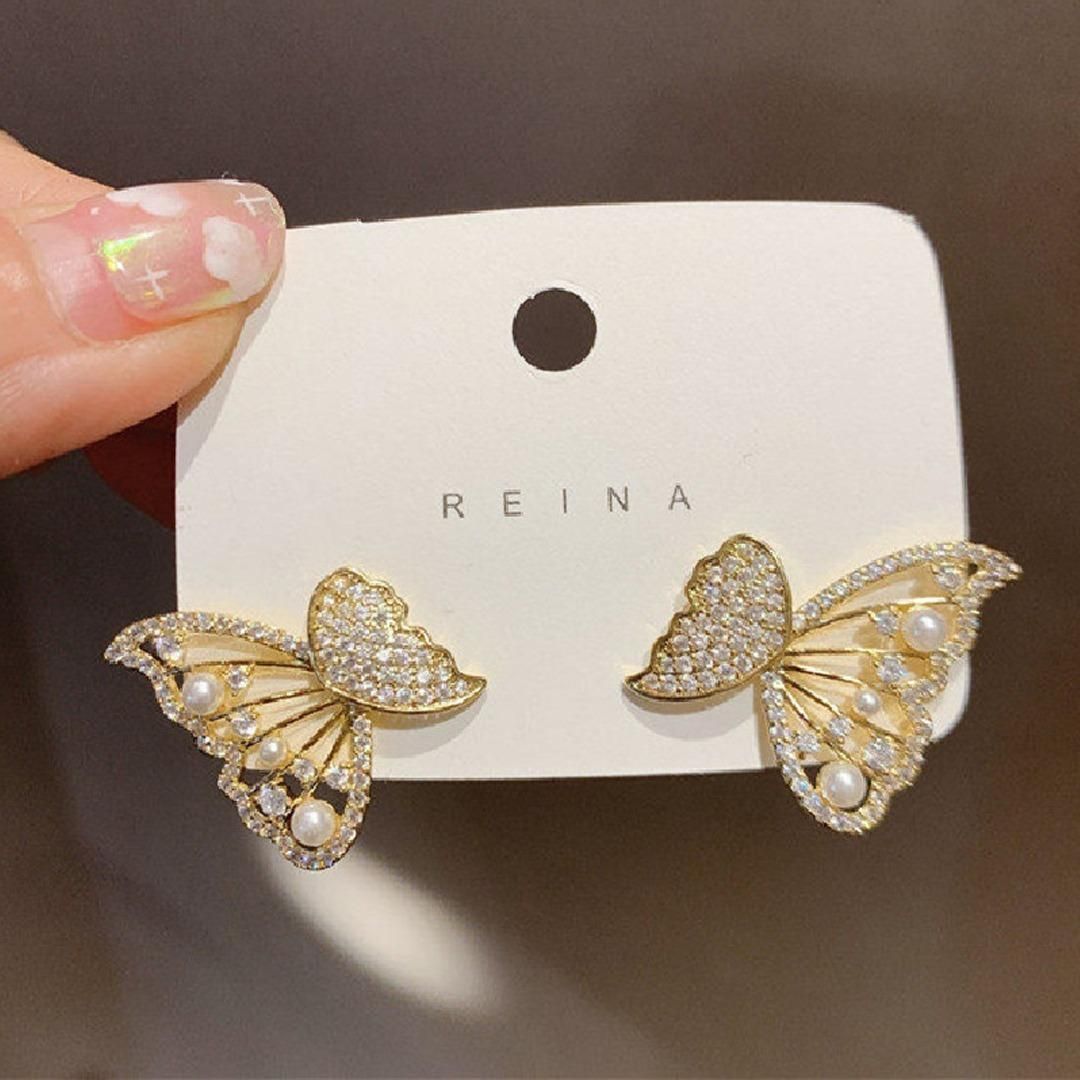A pair of gold butterfly-shaped earrings decorated with small gems and pearls, displayed on a card labeled 'REINA'.