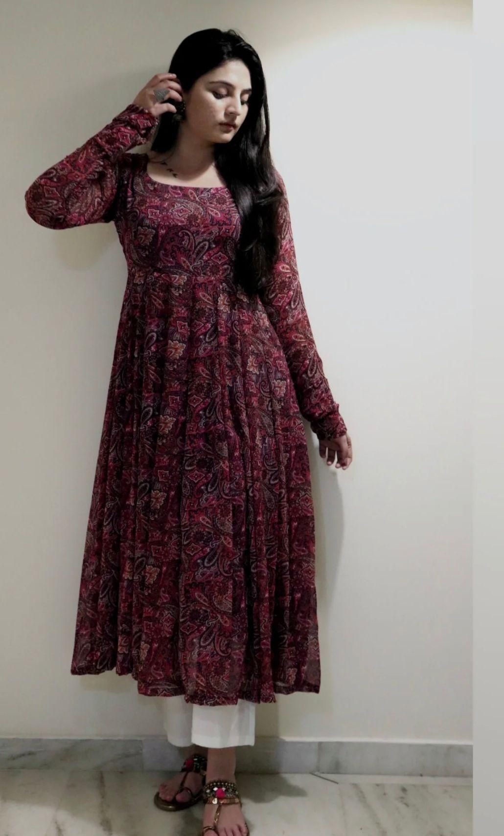 A woman wearing a long-sleeved red patterned dress with white pants and sandals, standing against a light-colored wall.