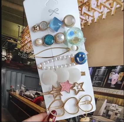 A hand holding a white display card with various colorful hair clips and decorative pins, including pearls, hearts, stars, and round shapes.