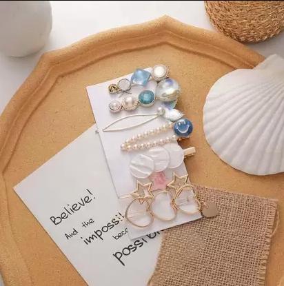 A display of various earrings and hairpins with pearl and blue accents, arranged on a card next to a large white seashell and motivational note.